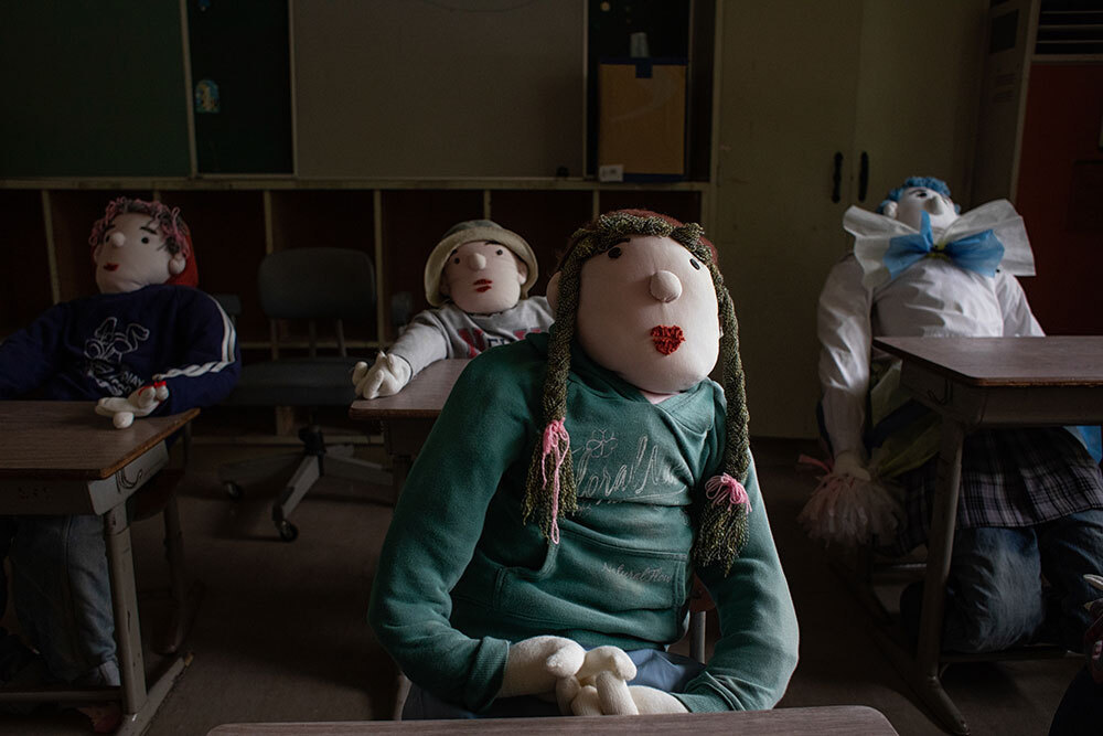 Life-sized dolls wearing clothes sit in school desks