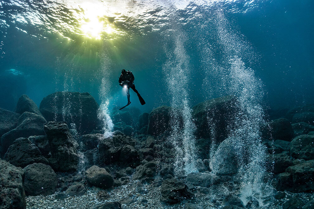 a person in a scuba suit underwater surrounded by bubbles being emitted by an underground magma chamber