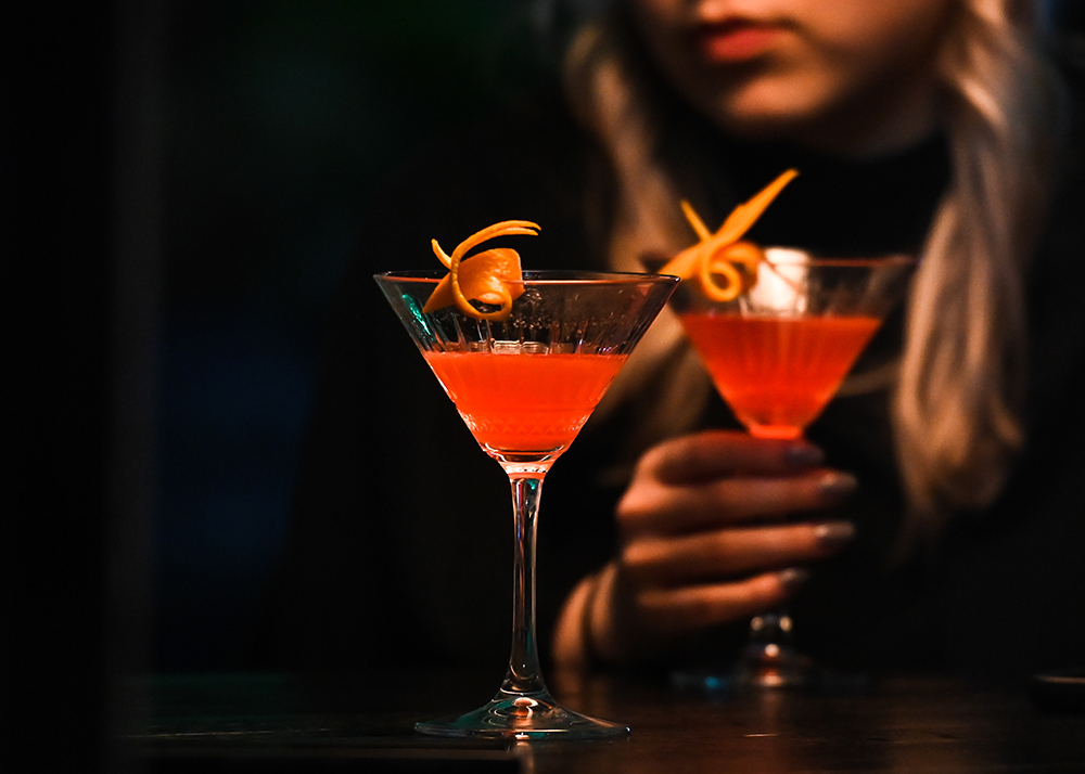 A woman drinking a pink cocktail from a martini glass, garnished with a twist of orange peel.