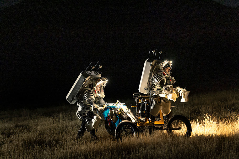 Two people wearing large, bulky suits walk in a dry field at night