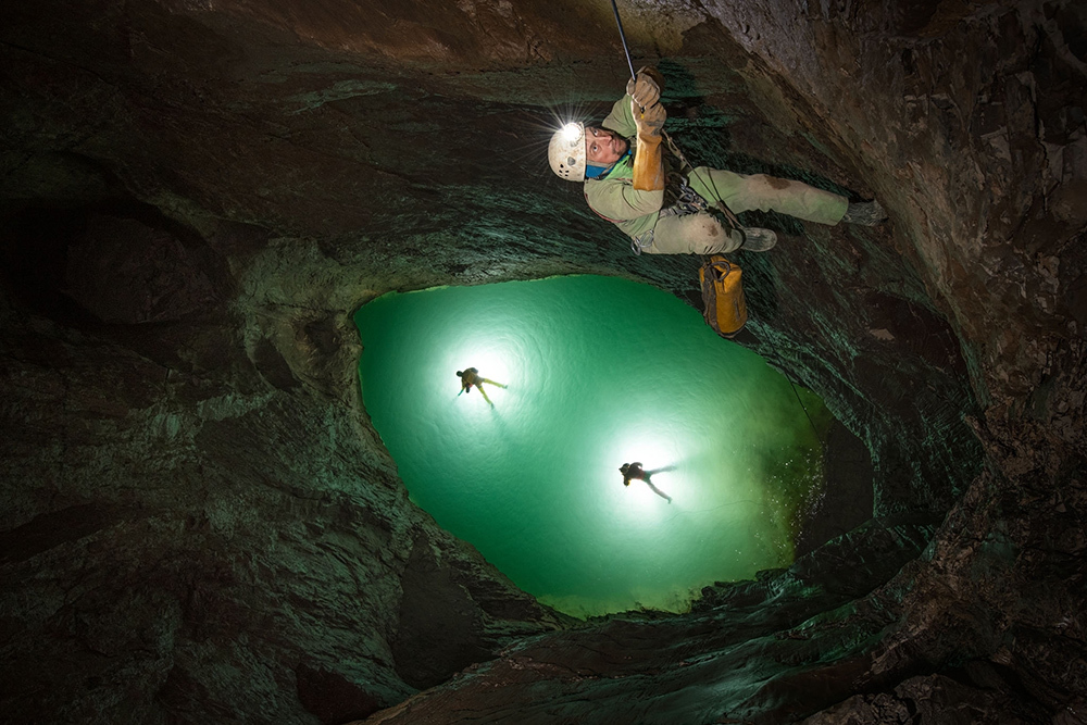 Man on cave wall and two people down in green water