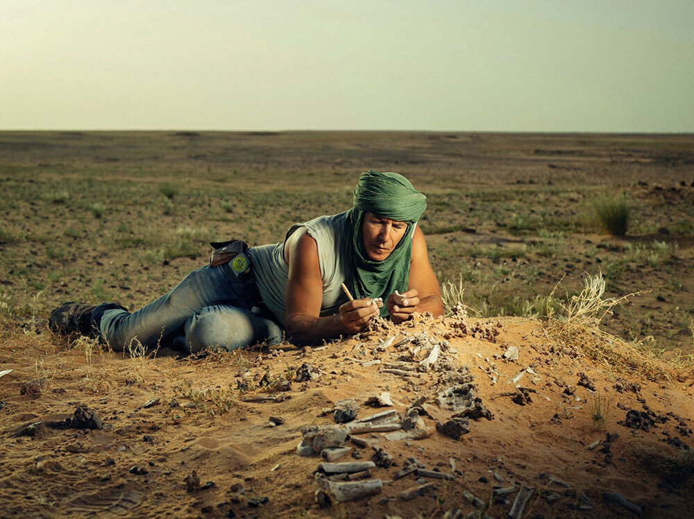 A man lies in the dirt looking at bones.