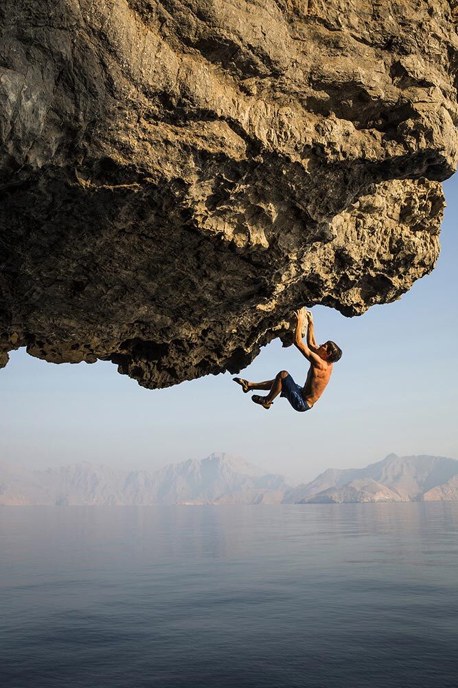 A sun-drenched climber, Alex Honnold, hangs from a rock overhang against a background of the ocean and distant mountains.