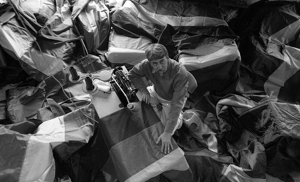 A top view of Gilbert Baker in black and white surrounded by flags at a sewing machine