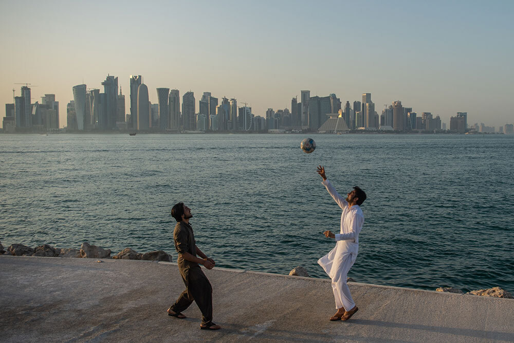 Two men toss a ball along the water with a city skyline in the background