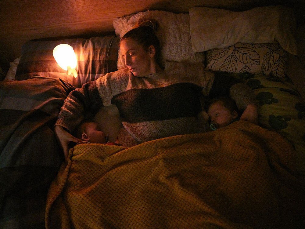 A mother lays down with her infant twins for a nap next to a lamp.