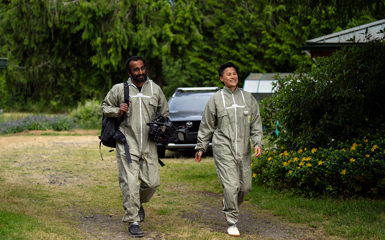 A photo of a man and woman in bee suits walking on a grassy path in an apiary.