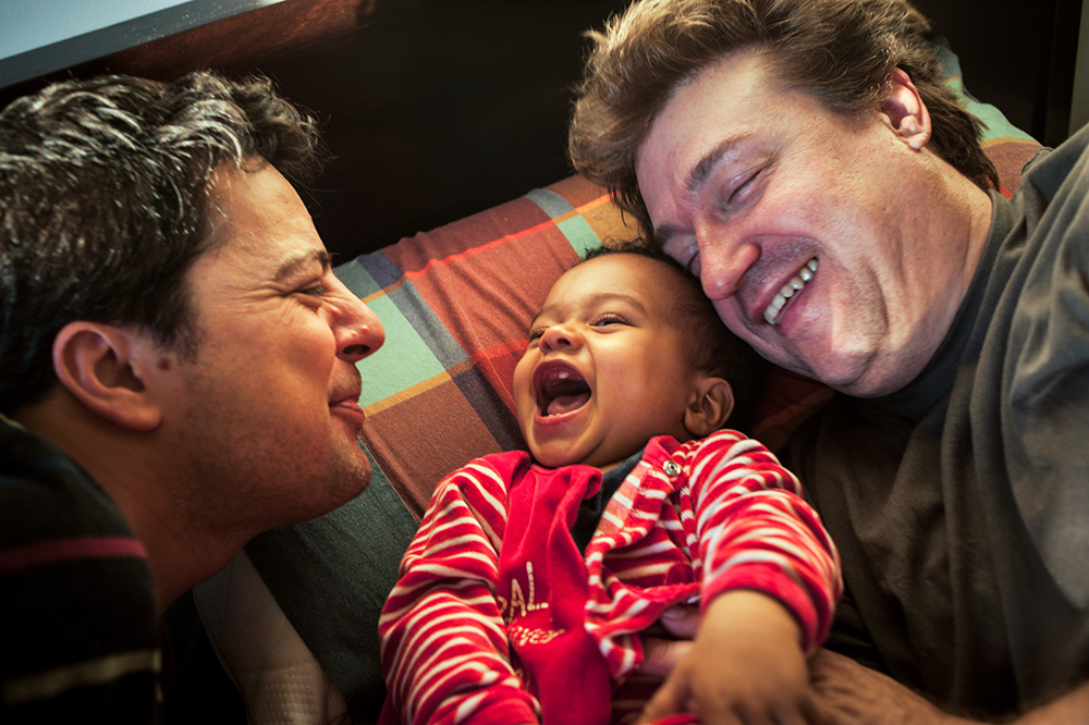 Two men embracing and cuddling their smiling, adopted baby.