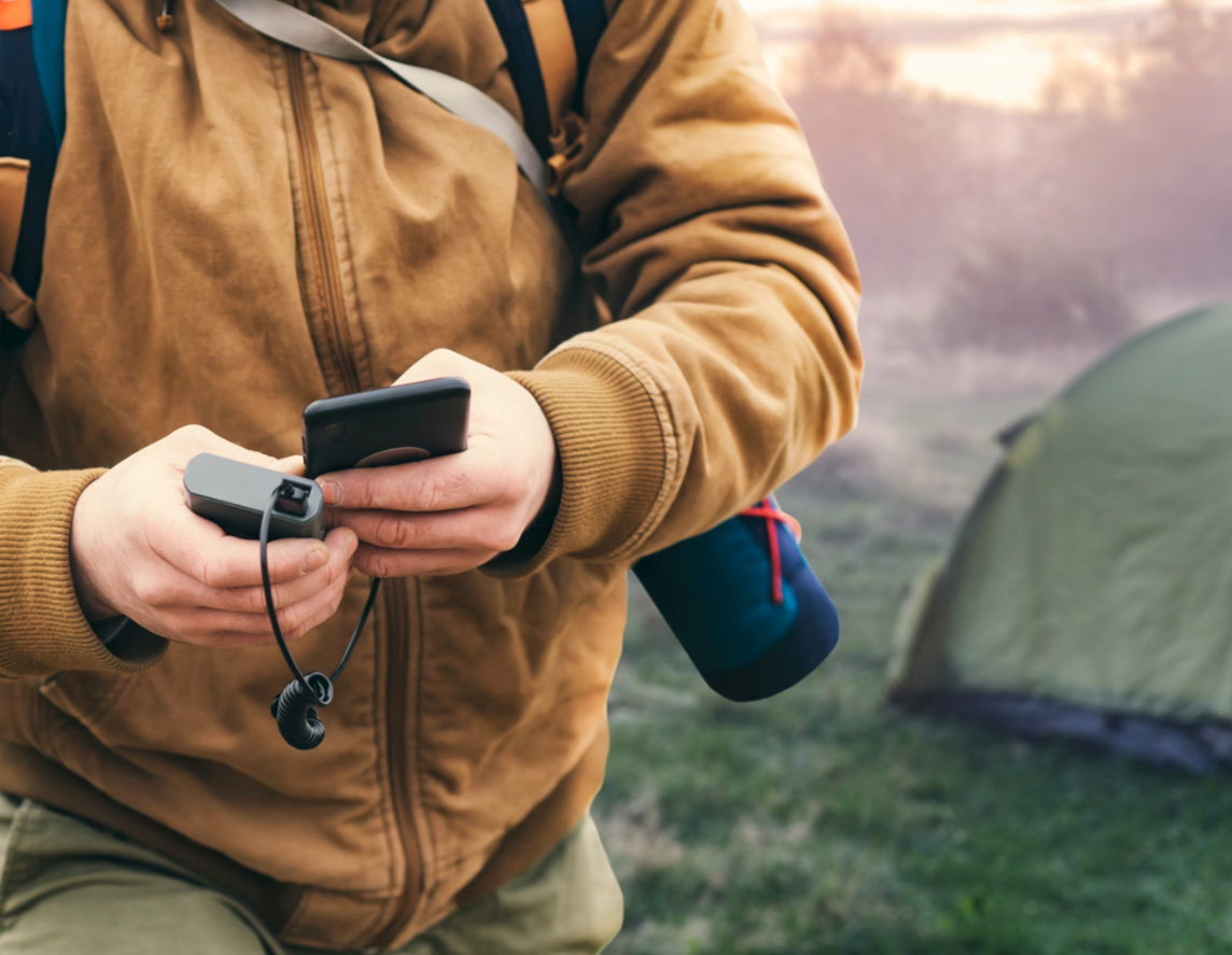 A close view of a man holding a smartphone in his hands as he charges it with a power bank, against the backdrop of a tourist tent in nature