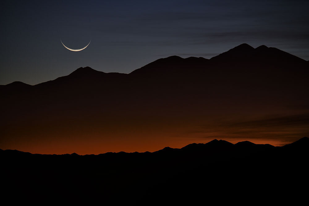 A sliver of a moon rises above a red horizon
