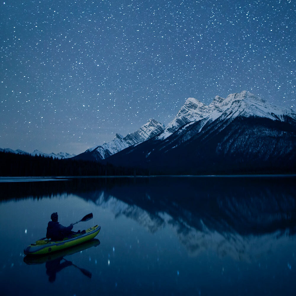 A person on a kayak sits on a still body of water with stars in the sky