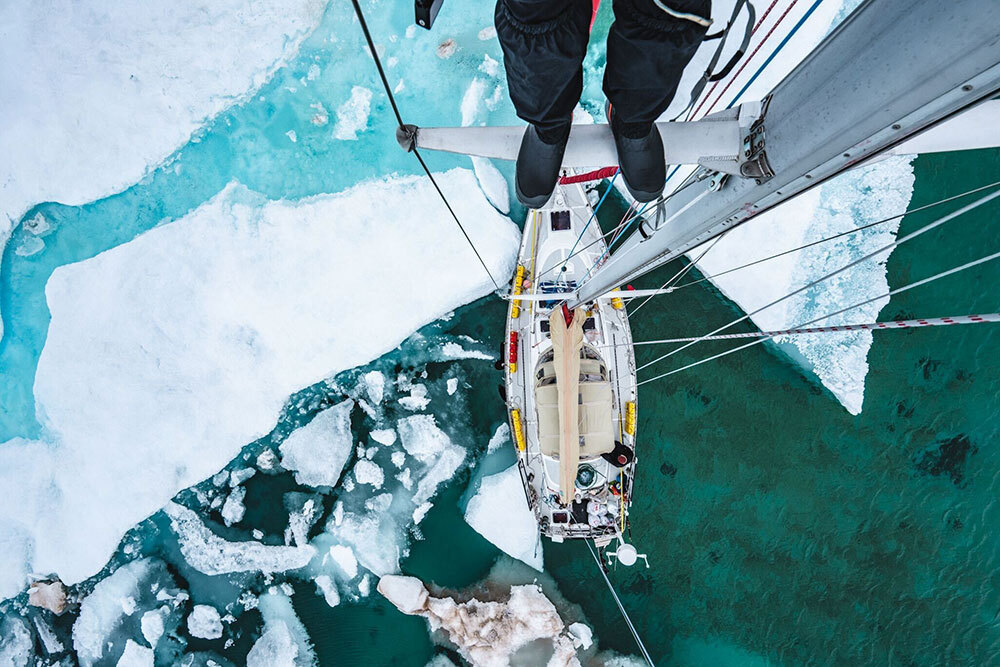 A person stands on a mast of a sailboat caught between ice