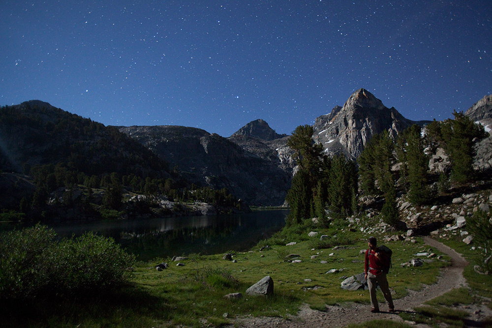 A hiker along a gravel path beneath a star-filled sky. Mountains stand tall in the background, illuminated by moonlight.