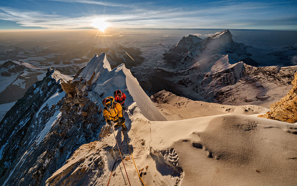 A picture of climbers on a mountain with the sun on the horizon in the background