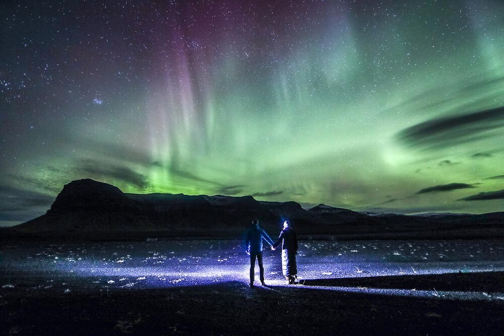 Two people hold hands while they watch the northern lights above a mountain range on Iceland's south coast.