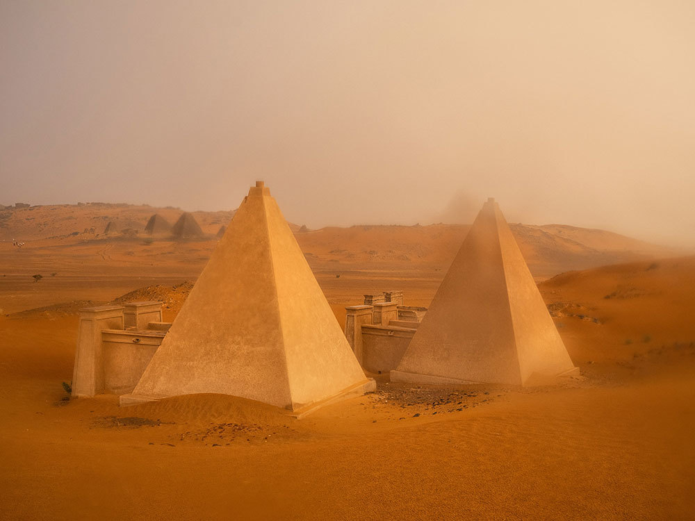 A picture two pyramids in the desert with a darkened, hazy brown sky