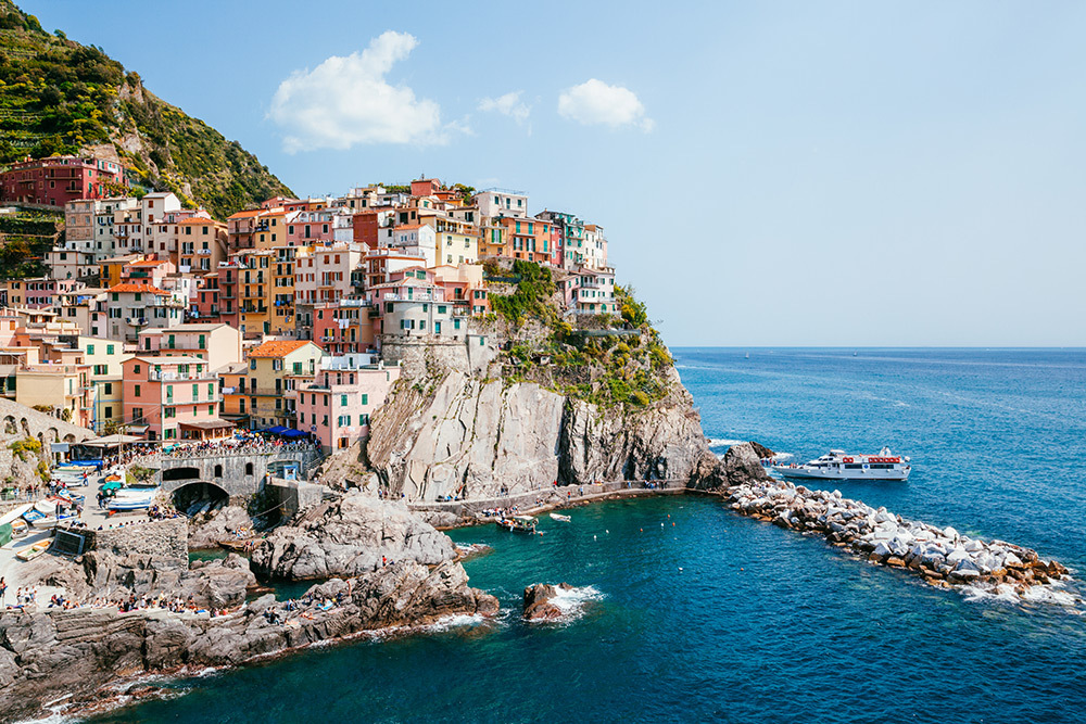 A view of colorful houses atop a cliff in Italy. Below the sea is calm, and a boat is coming in from the sea