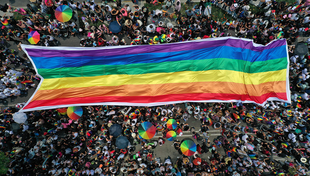 A top view of people in a pride parade with a large pride flag in the center and people walking with pride umbrellas around it.