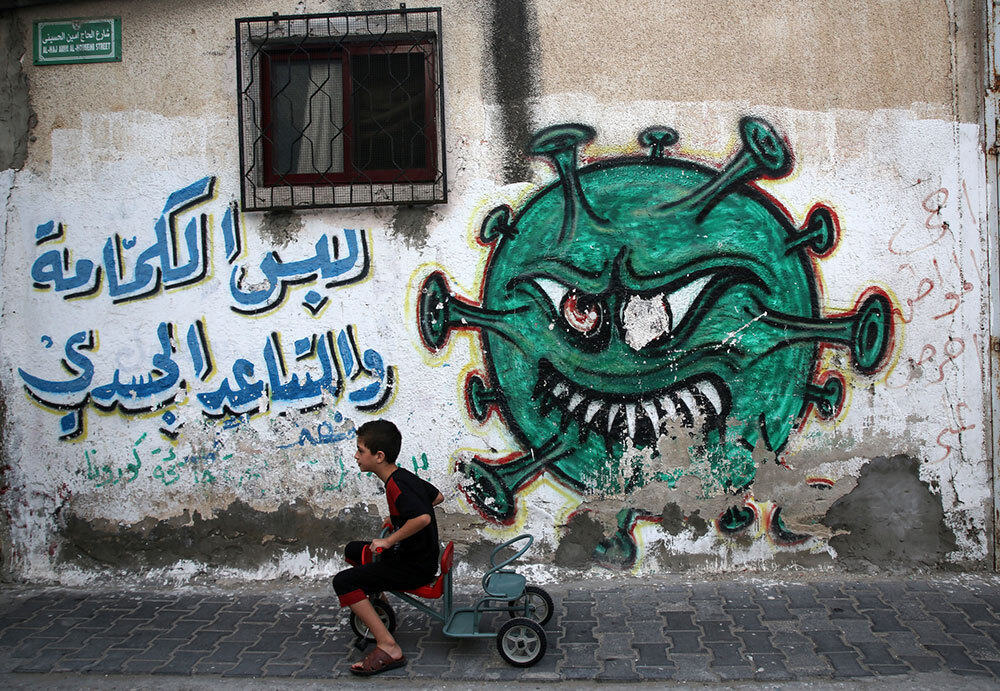 A kid scoots a tricycle past a graffiti image of a COVID-19 virus