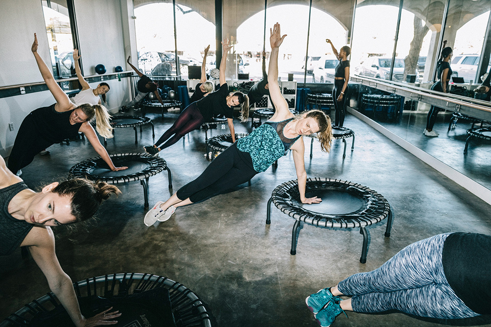 A class of women all using one arm to support their weight as they lean on small trampolines.