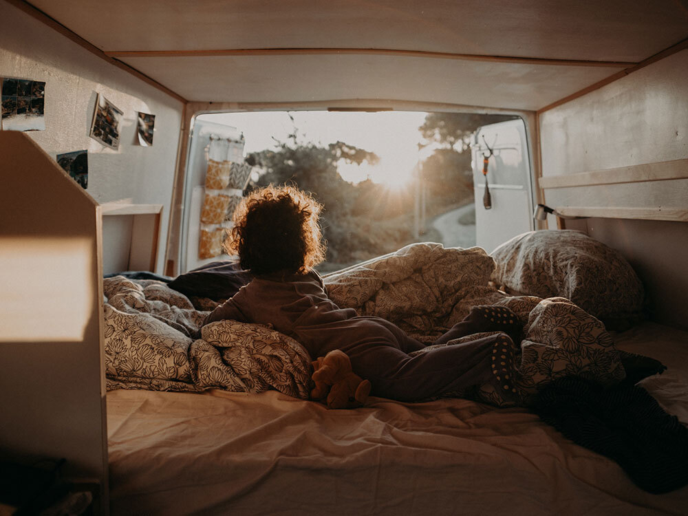 A toddler slowly wakes up from his night in a van. He watches the sunrise through the open back doors of the van.