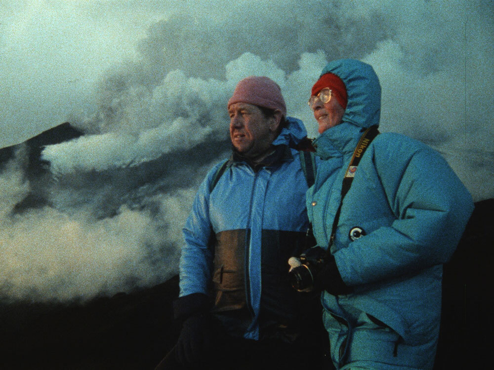 Katia and Maurice Krafft gaze at a volcano in the distance as plumes of ash, steam, and gasses billow behind them.