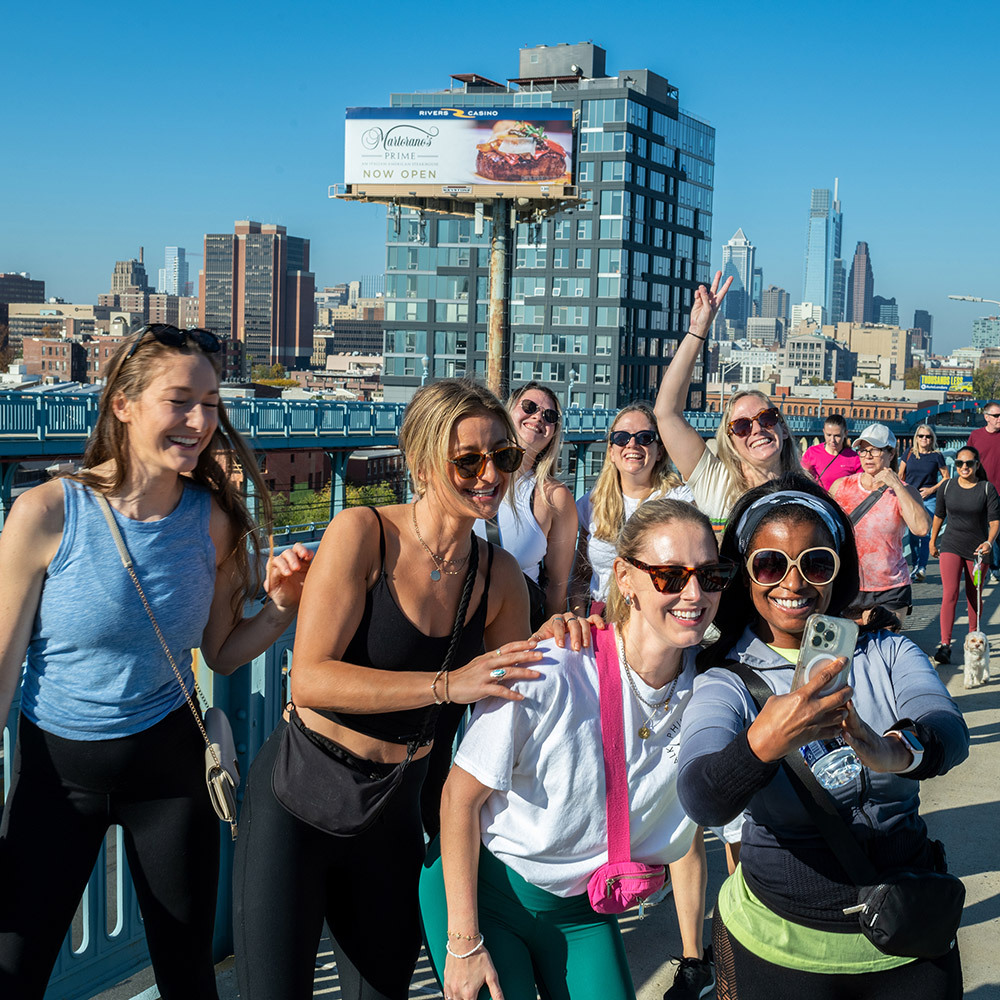 A crowd of people walk over a city bridge. Seven women at the front pose for a group selfie together