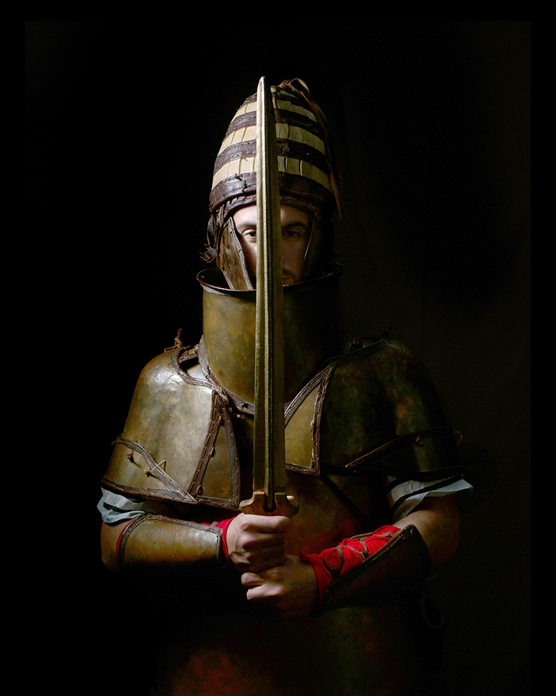 A person poses for a studio portrait with a sword and helmet. They appear to just be coming out of the shadow.