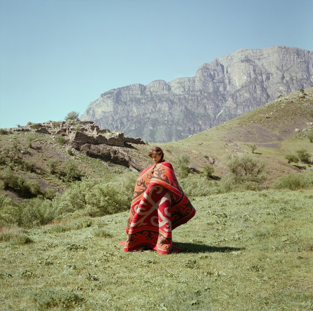 A woman drapes a carpet over herself in a field with a ruined castle and mountains in the background.