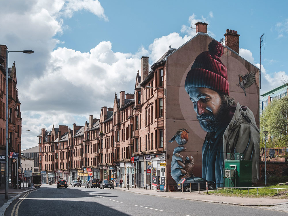 A mural of a man on the side of a row of buildings. The man is wearing a beanie and a bird is perched on his finger