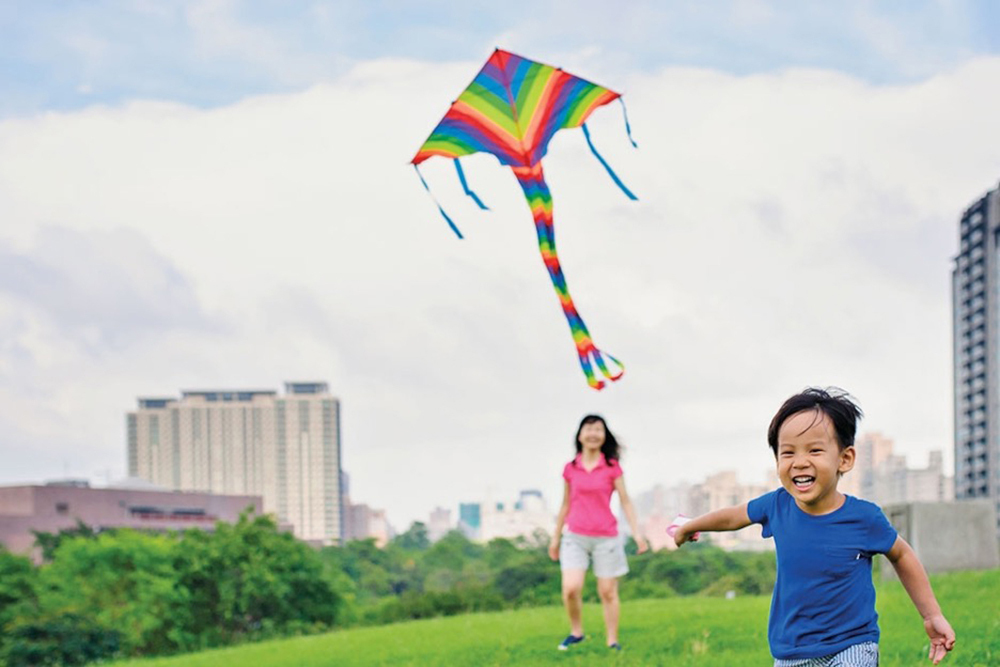 A child flies a kite in the air with the parent and city in the background.