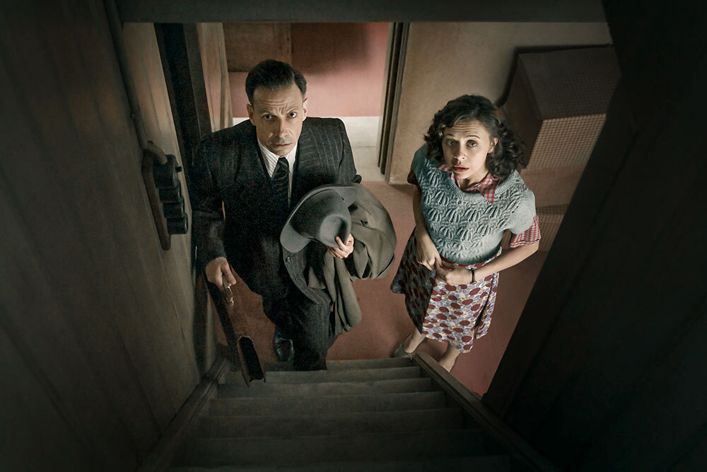 a still image from the limited series show 'A Small Light' showing a man and a woman at the bottom of a staircase looking up