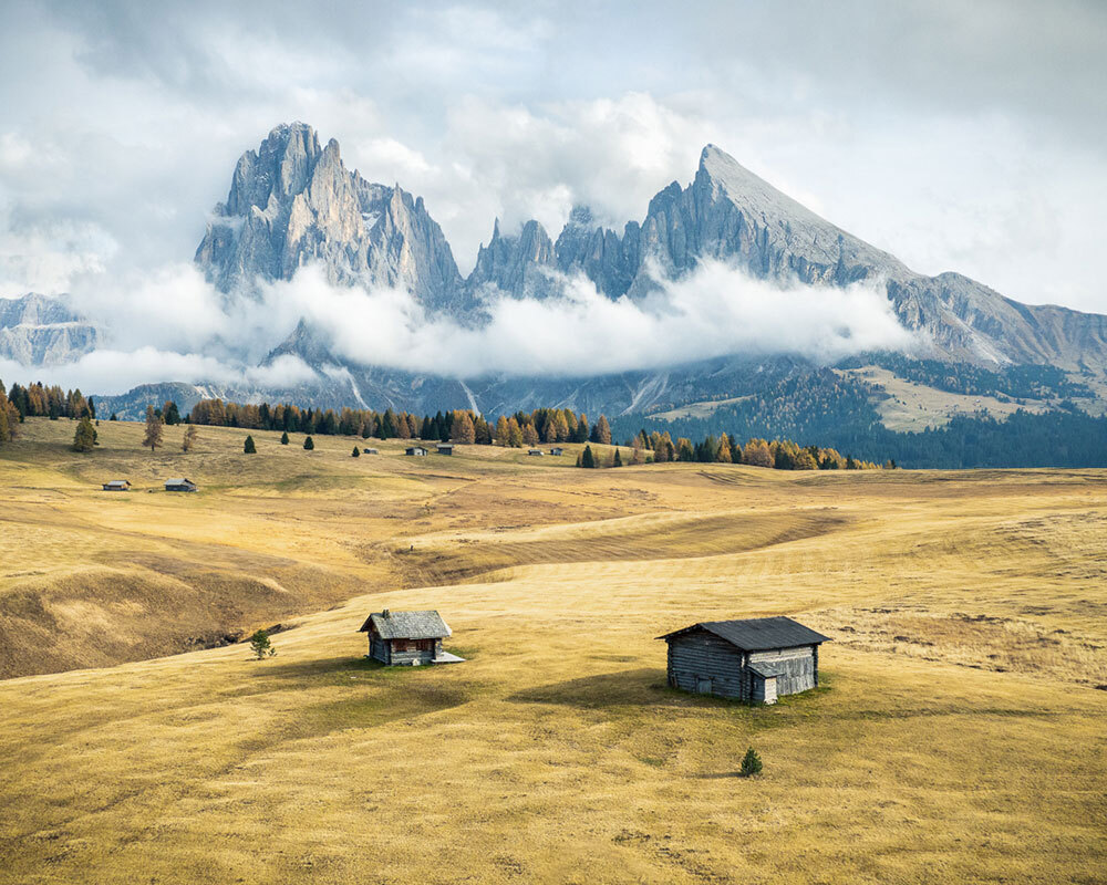 Two buildings stand in a golden field with mountains in the background