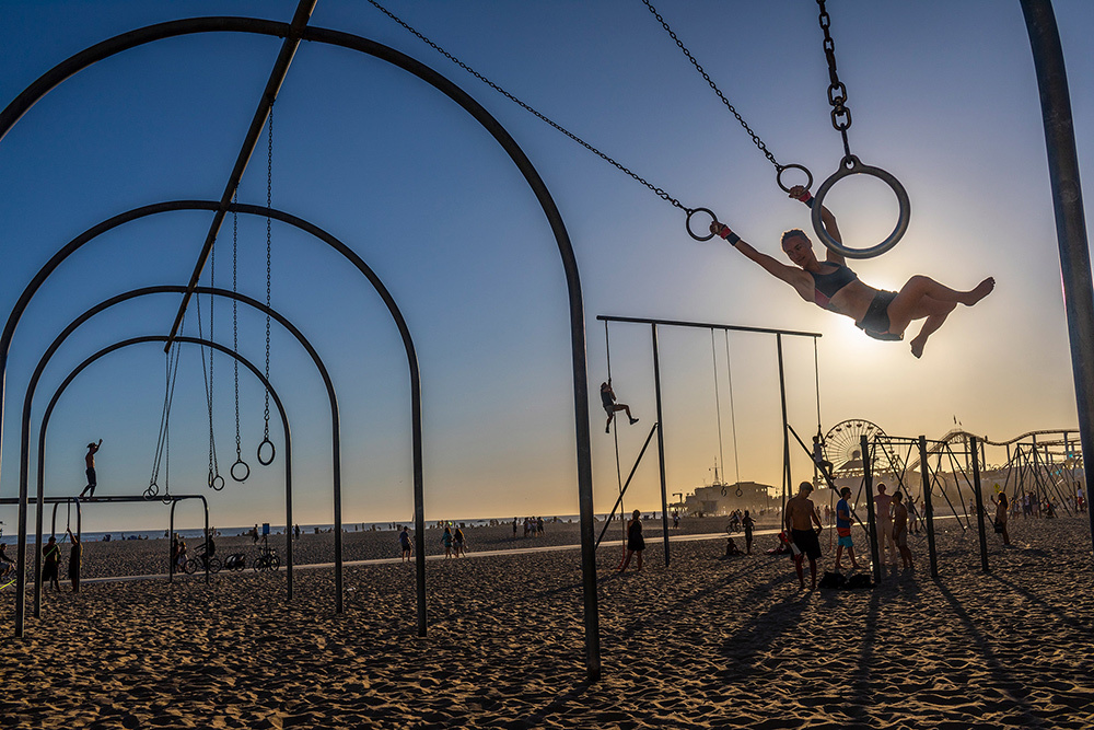 People are silhouetted on a back, a person swings from rings and others use various equipment in the distance.