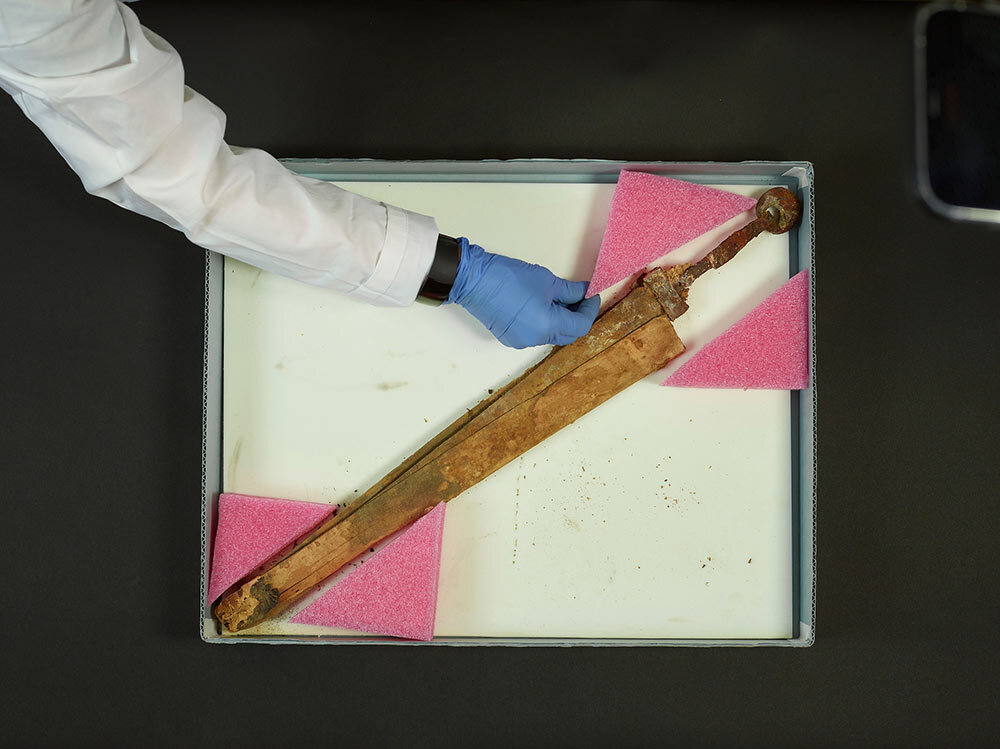 A conservator adjusts a foam wedge protecting the distinctive iron hilt of a Roman ring-pommel sword.