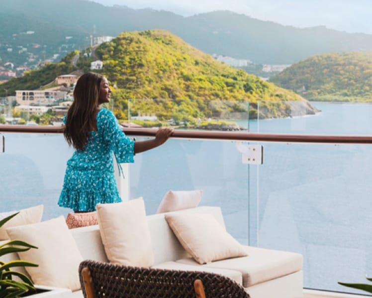 A woman on the deck of a cruise ship gazes serenely at the water and lush green islands.