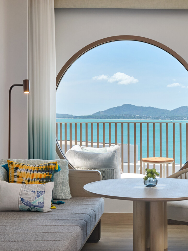 A hotel room with a large circular window with a view of a balcony, the ocean, and a mountain range.