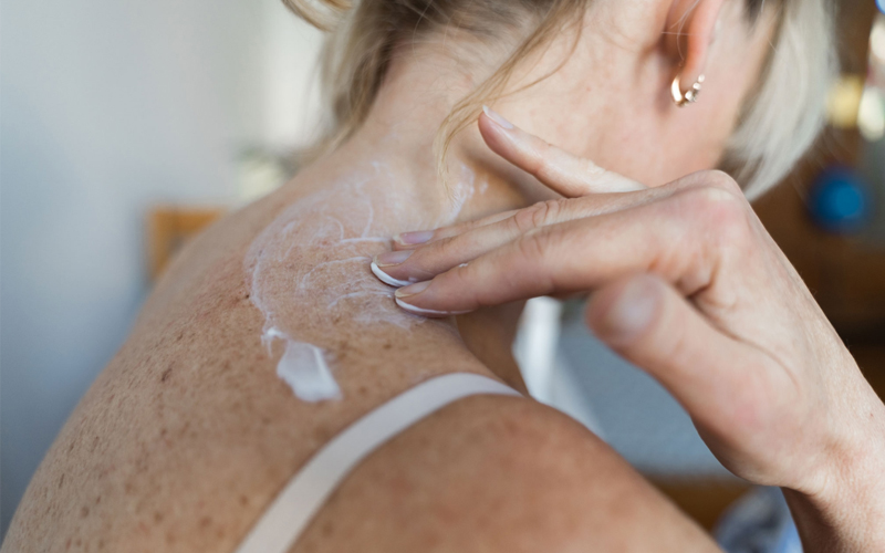 A woman rubs cream onto her upper back.