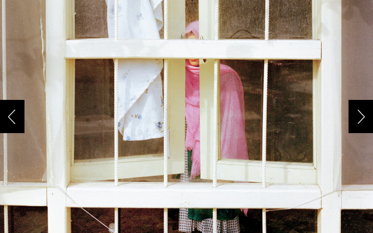 A girl in pink is photographed standing beside a window, her face obscured. 