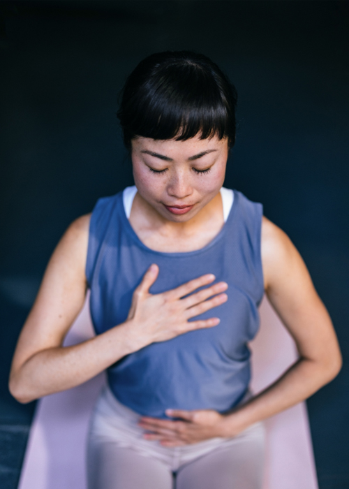 An East Asian woman in workout clothes has her eyes closed and her right hand over her heart and left over her stomach.
