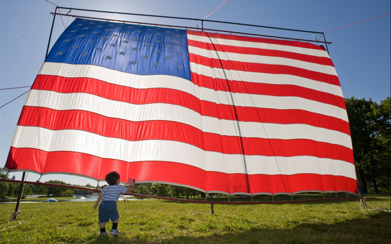 A toddler walks towards the front of a large American flag propped up on a lawn.