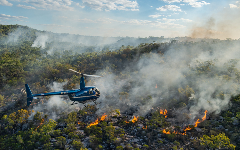 A helicopter flies over a section of a large forest on fire. 