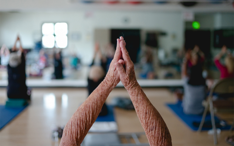 The wrinkled arms of an older woman as she brings her palms together during a stretch in a yoga class.