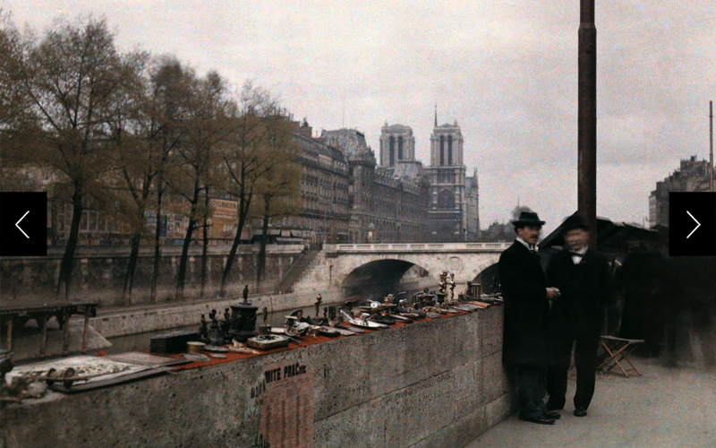 Two men stand next to trinkets for sale on the banks of the Seine in the 1920s with Notre Dame in the background. 