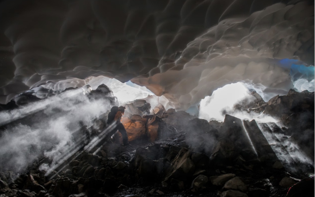 An expedition member is walking through a cave entrance with beams of light streaming in through fog. 