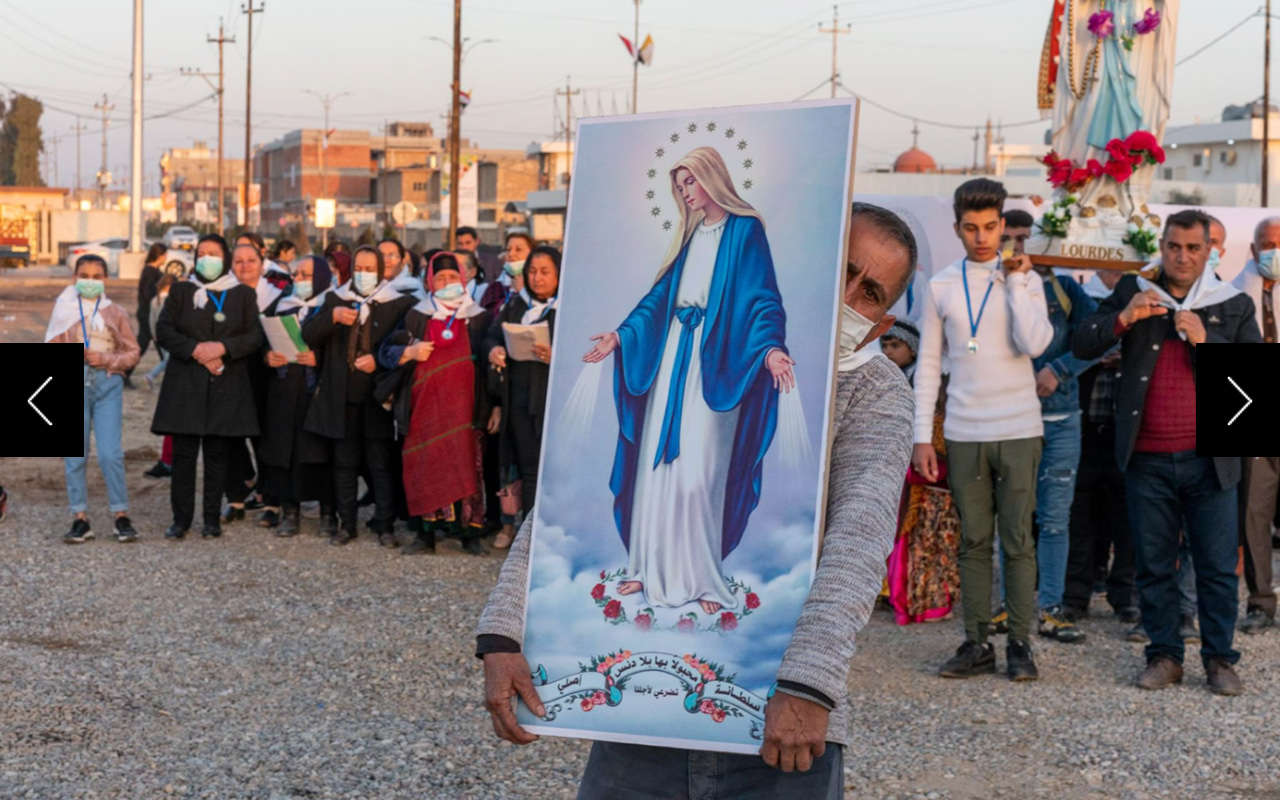 A man holds a large illustration of the Virgin Mary while dozens of other people march behind him. 