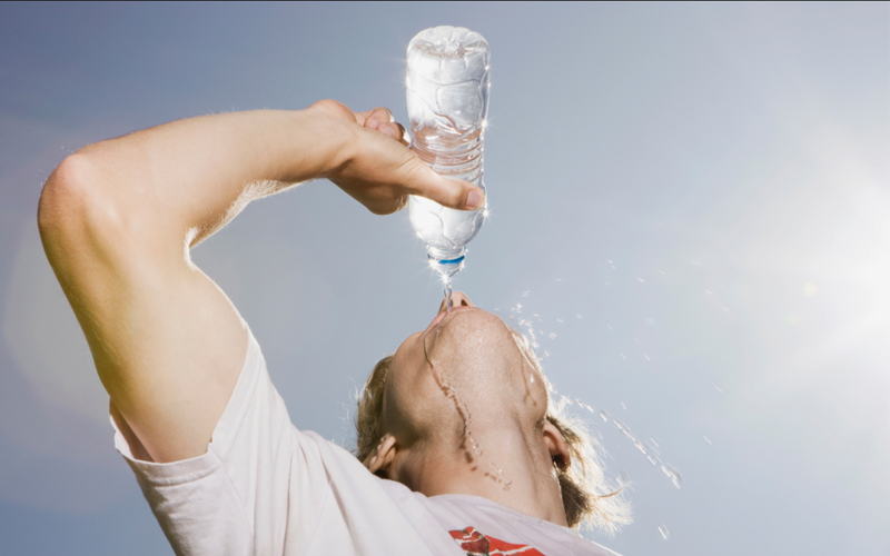 A person leans their head back as they drink from a plastic bottle of water. 